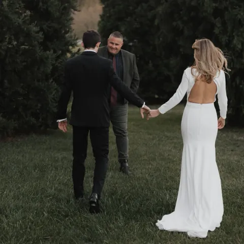 A couple walking hand in hand across grass towards their celebrant, who is smiling as they come up to him.